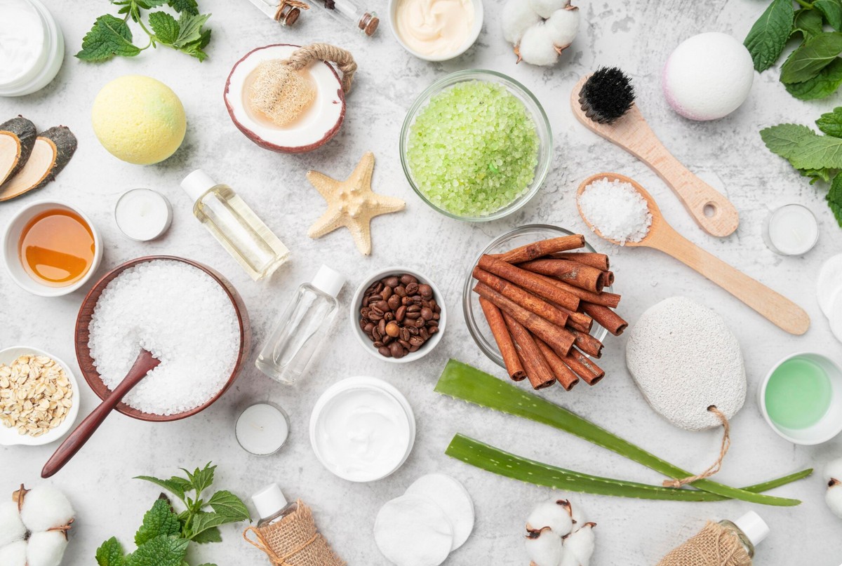 Flat lay of raw skincare ingredients including sea salt, aloe, coconut, oats, cinnamon and botanical oils on a pale stone surface