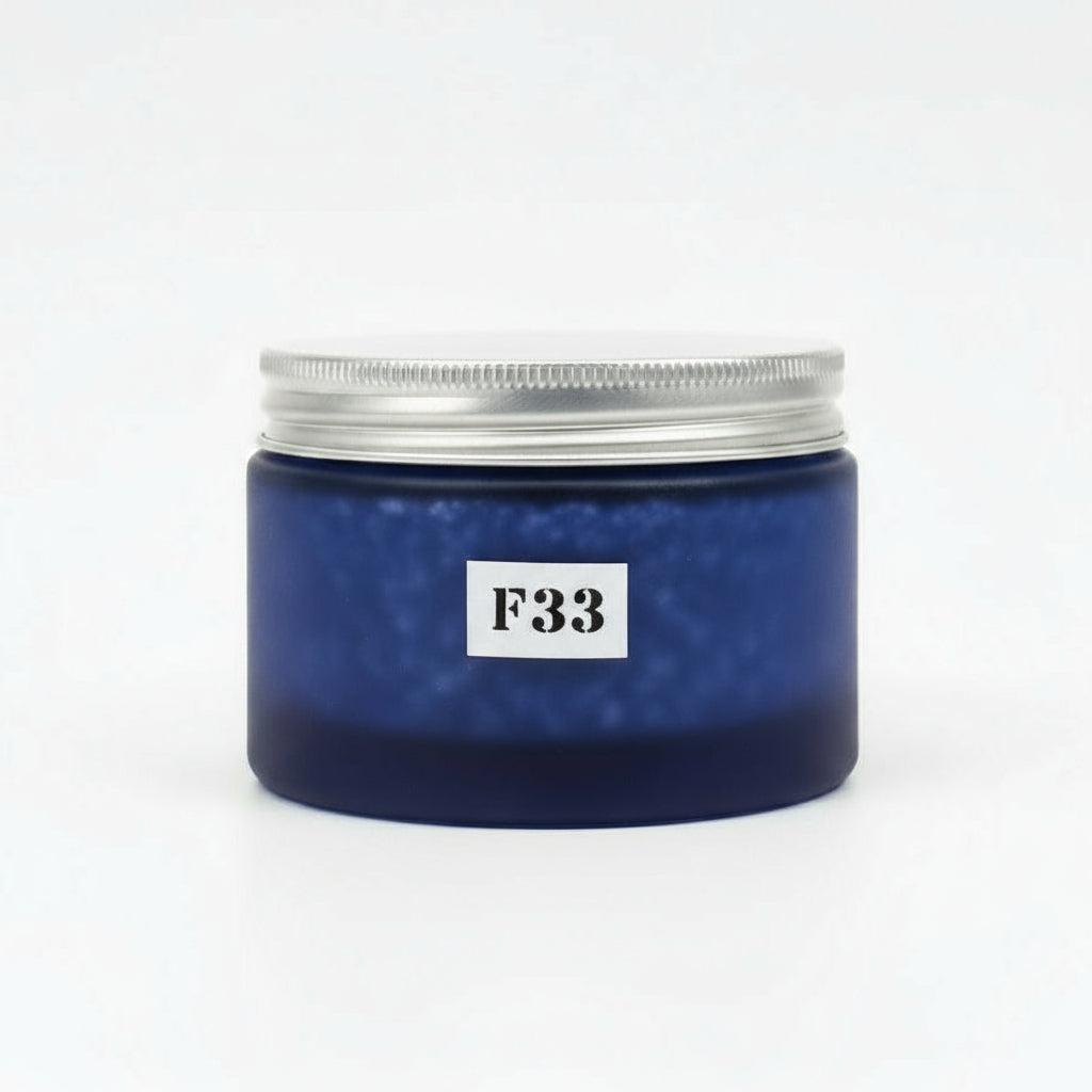 A jar of body scrub with a blue label reading 'F33'. The jar is placed on a wooden surface with green foliage and pink flowers in the background.