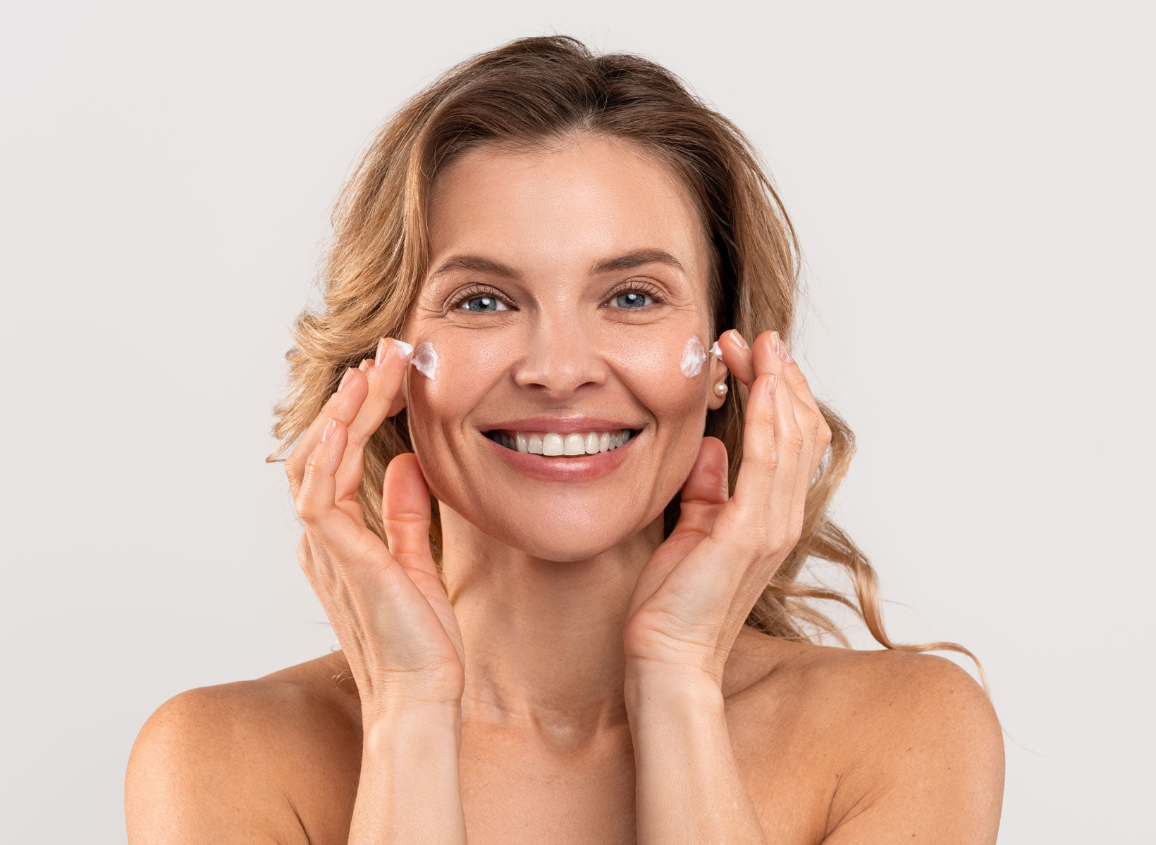 A smiling woman facing straight in puts skin cream on both cheeks of her face